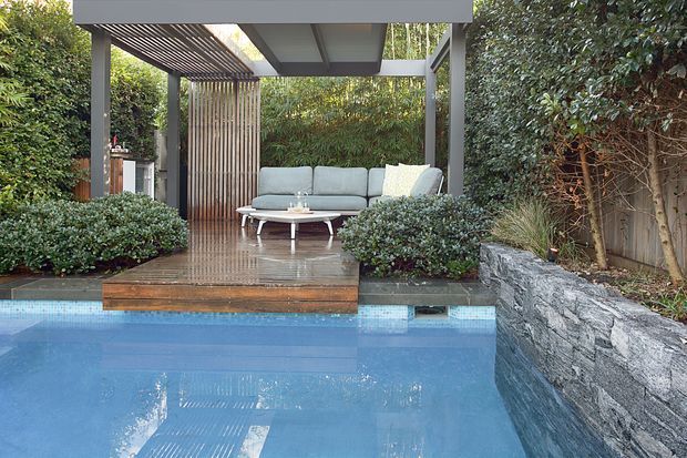 A landscaped outdoor pool area with a raised timber deck and a sheltered lounge pavilion. The pavilion has a flat roof, dark structural posts, a slatted timber pergola, vertical timber screening, and a grey outdoor sofa arranged around a low coffee table. Dense planting surrounds the space, while a rough stacked-stone retaining wall borders the pool.