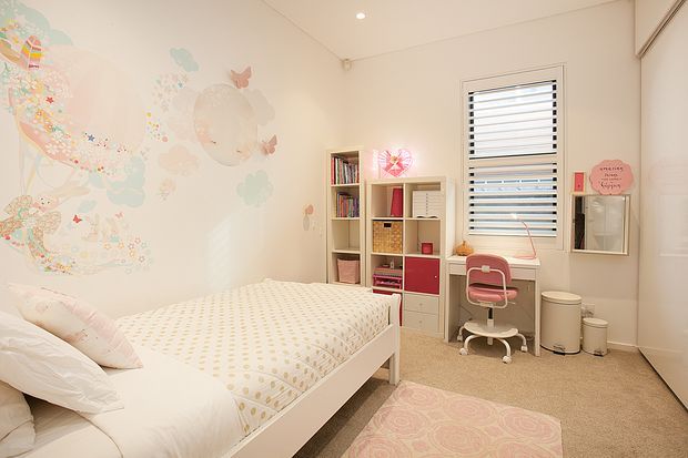 Bright, softly styled children's bedroom with a white bed, pastel patterned bedding, whimsical mural decals, built-in shelving and storage, a small pink desk chair, and a window with horizontal blinds. The room has a warm neutral carpet, white walls and joinery, and playful pastel accents.
