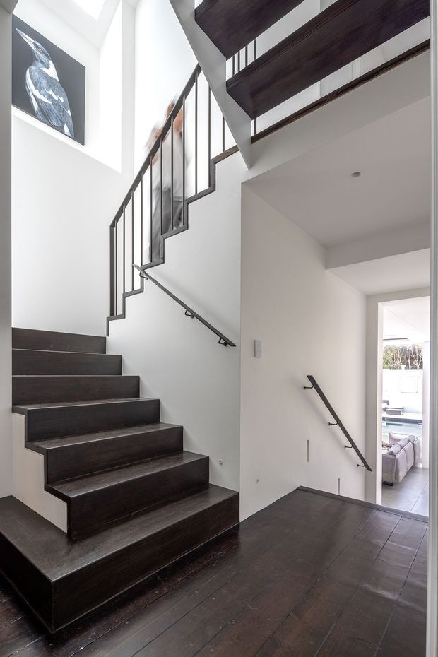 A bright, contemporary stairway and landing with dark timber stair treads, risers and flooring contrasted against white plaster walls. Slim black metal balustrades frame the open staircase, which rises beside a tall void with strong natural light from above. A dark timber mezzanine or upper-level edge is visible overhead, and a monochrome bird artwork hangs on the upper wall. The landing looks through to a bright living area beyond.