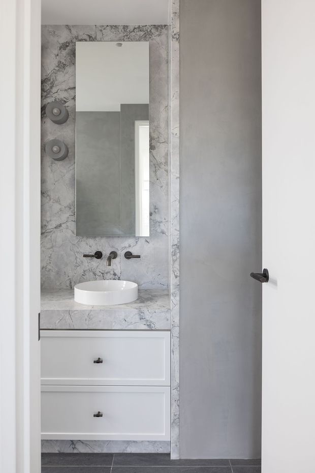 A compact contemporary bathroom viewed through an open doorway. The vanity has white shaker-style drawers, a white vessel basin, dark wall-mounted tapware, and a grey-white marble-patterned benchtop and wall surround. A tall rectangular mirror is mounted above the basin, with two round wall lights to its left. The floor is finished in dark grey tiles, and the partially open grey door occupies the right side of the image.