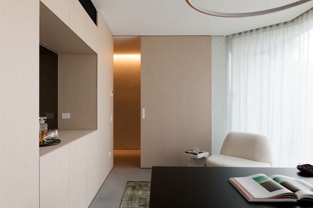 A refined, compact contemporary interior featuring pale wood built-in cabinetry and a recessed display niche, a dark stone or composite tabletop in the foreground, a softly illuminated bronze-toned passage, a cream lounge chair, and full-height sheer curtains. A curved ceiling detail with integrated trim adds architectural interest, while the restrained palette creates a calm, luxurious workspace or living area.