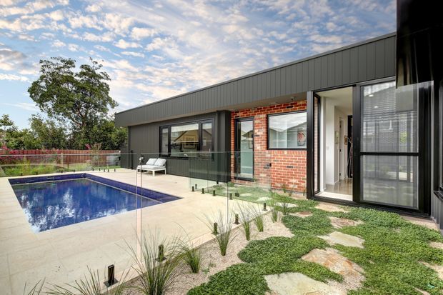 Single-storey contemporary residence viewed from the landscaped pool terrace. The home has a long low-pitched dark metal roof, dark-framed glazing, a red brick feature wall, and a partially enclosed outdoor entertaining area beside a rectangular swimming pool. Pale paving, clipped groundcover, ornamental grasses, and stepping stones form the surrounding landscape.