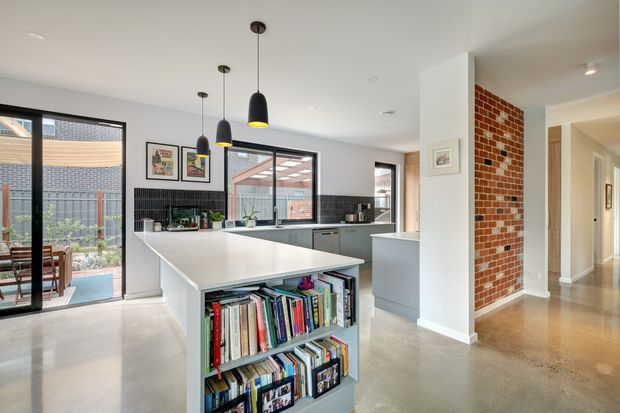 Bright open-plan kitchen viewed from the living area, featuring a large white island with integrated open shelving, grey cabinetry, black tiled splashback, pendant lighting, polished concrete flooring, and exposed red brick detailing. Full-height glazing opens toward a small outdoor courtyard or patio, while a hallway is visible beyond the kitchen.