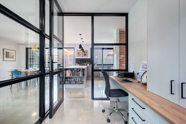 Bright contemporary home office viewed through black-framed internal glass partitions, with a timber desk, ergonomic office chair, white built-in cabinetry, polished concrete flooring, exposed brick detailing, and a visible open-plan dining/living area beyond.