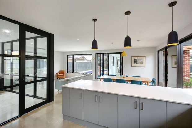 A bright open-plan kitchen and dining area with a large grey island, white benchtop, black-framed glazed doors and windows, pendant lighting, and a timber dining table. The space opens toward a relaxed living area and features polished concrete flooring with a neutral contemporary palette.