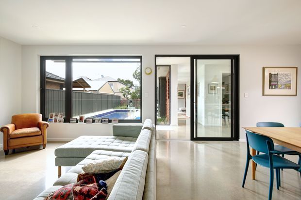 Bright contemporary open-plan living and dining area with polished light flooring, a large sectional sofa, armchair, timber dining table, blue dining chairs, and black-framed glazing and internal doors. The room opens toward a hallway and other interior spaces, with a view to a backyard and pool through the window.