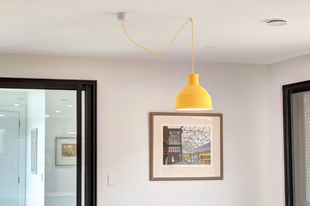 Bright contemporary interior view of a white-painted room with a vivid yellow pendant light suspended from the ceiling, a framed architectural artwork, and dark-framed glazed doorways on either side. A bathroom or wet area is visible through the left opening, while the main space appears to function as a circulation or entry area.