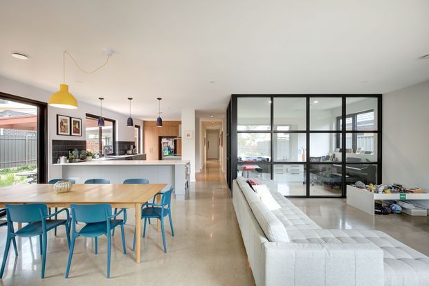 Open-plan contemporary home interior combining a dining area, kitchen and living room. The space features polished concrete flooring, a white kitchen with island, timber dining table, colourful dining chairs, pendant lighting, and a glass-and-black-metal framed home office or study enclosure. Large glazing provides views to an outdoor area.