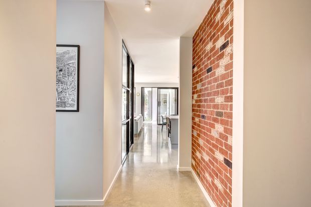 A bright, narrow interior hallway with polished concrete flooring, white walls and ceiling, black-framed glazed openings, and a prominent exposed red brick feature wall. A framed architectural print hangs on the left, and the corridor leads toward a glazed doorway and a glimpse of a furnished room beyond.