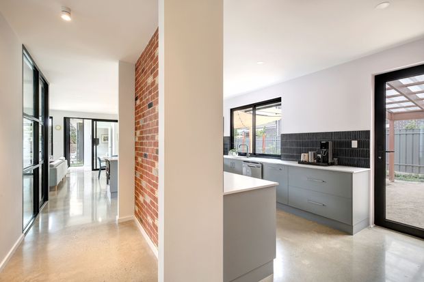 Bright contemporary open-plan interior showing a polished concrete-floored hallway and kitchen. The kitchen has muted grey-blue cabinetry, a dark vertically ribbed tile splashback, pale benchtops, black-framed glazing and doors, and an exposed red brick feature wall along the hallway. Large windows and glazed doors provide abundant natural light and views to an outdoor area.