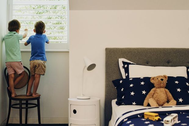 A children's bedroom with two young children looking through white louvred window shutters. The room features a single bed with a dark upholstered button-tufted headboard, navy star-patterned bedding, a teddy bear, toy vehicles, and a small white bedside table with a modern lamp. The palette is soft and neutral with playful navy accents.