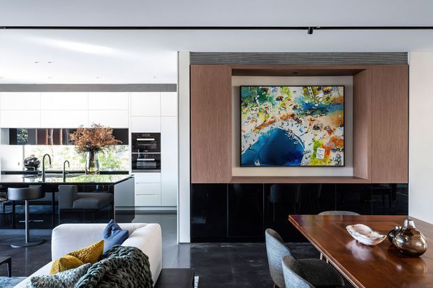 Open-plan contemporary living and dining area connected to a sleek kitchen. The room features dark stone-look flooring, white and timber-toned cabinetry, a black built-in media unit with a large colourful artwork, and a polished timber dining table with upholstered chairs. A sofa and kitchen island are visible, creating a refined, modern entertaining space.