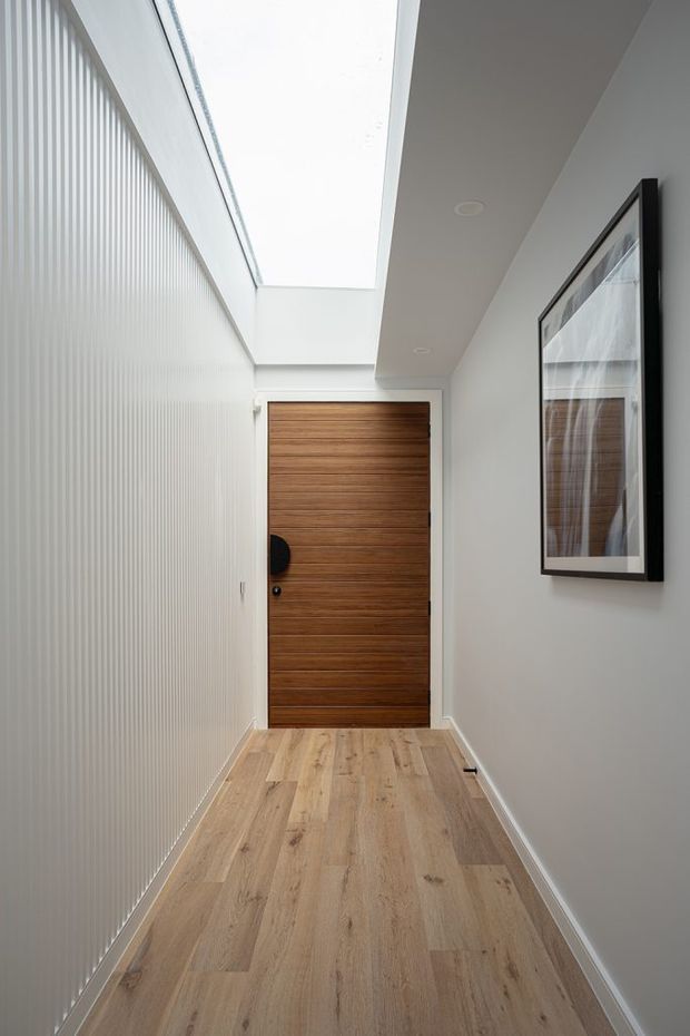 A narrow, bright residential hallway leading to a horizontal timber front door. The corridor has pale timber-look flooring, a white vertically ribbed wall finish on the left, smooth white walls and ceiling, recessed downlights, a large linear skylight, and a black-framed artwork on the right wall.