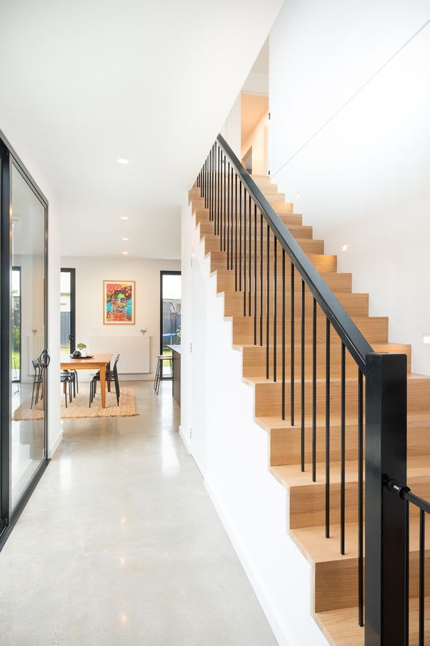 Bright contemporary interior view focused on an open staircase with warm timber treads and risers, slim black metal vertical balusters, and a dark handrail. A polished light concrete hallway floor leads toward a dining area with timber table and chairs, white walls, large internal glazing, and minimalist recessed lighting.
