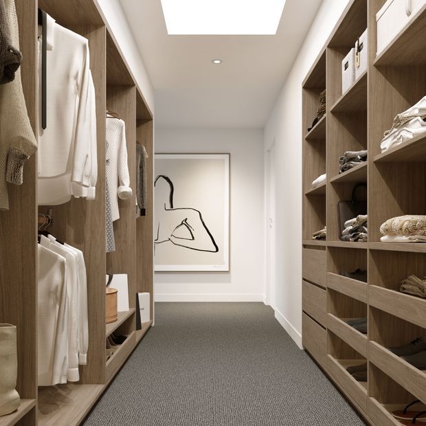 A well-organised walk-in wardrobe with full-height open timber shelving and hanging sections on both sides, displaying folded and hanging clothing. The space has a soft grey carpet, white walls and ceiling, integrated recessed lighting, and a large rectangular skylight or ceiling light. A minimalist framed line artwork is positioned at the end of the corridor, creating a refined, warm and highly functional dressing area.