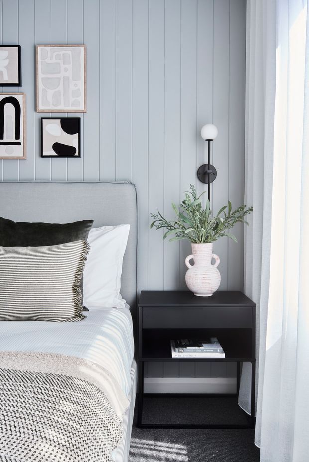 A softly styled bedroom corner with a light grey upholstered bed, layered neutral bedding and cushions, vertical pale grey wall panelling, framed abstract artwork, a black open nightstand, and a slim black wall-mounted globe sconce. A large sheer-curtained window brings in bright natural light, while a ceramic vase with greenery decorates the bedside table.