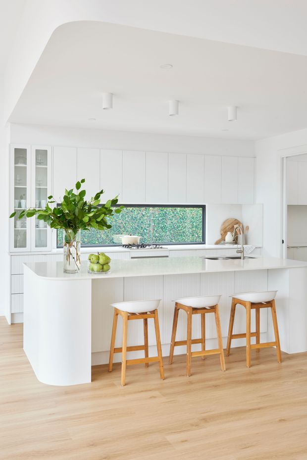 Bright, predominantly white contemporary kitchen with a large curved-edge island and integrated breakfast bar seating for three. The space features white full-height cabinetry, glass-front display cupboards, a pale stone-look benchtop, timber flooring, recessed and cylindrical ceiling lights, and a long horizontal window overlooking greenery. Natural timber stools and a leafy arrangement add warmth.