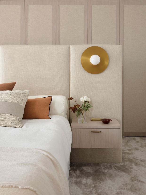 A softly styled bedroom corner with a partially visible bed, upholstered cream headboard, layered neutral and rust-toned cushions, and a matching bedside table. The room features warm beige wall panelling, a gold circular wall light, pale timber joinery, and a plush grey-beige carpet, creating a refined, calm, neutral-toned interior.