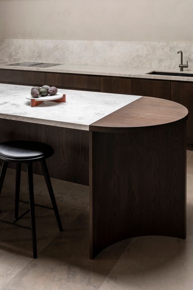 A sophisticated modern kitchen featuring dark wood cabinetry and a sculptural island. The island has a pale veined stone benchtop with an integrated rounded dark timber extension, paired with a black bar stool. Matching dark cabinetry runs along the back wall beneath a pale stone splashback, with a dark metal tap and inset sink. The space has a warm, moody, refined atmosphere and understated natural materials.