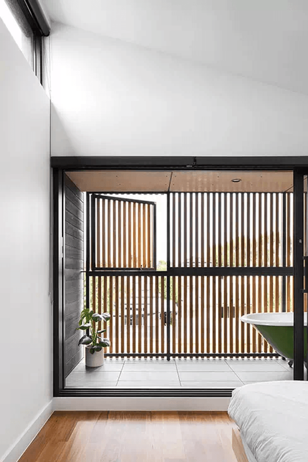 A bright, minimalist bedroom viewed toward a glazed balcony or courtyard. The room has white walls, warm timber flooring, black-framed full-height glazing, and a partially visible bed. Outside, a tiled terrace is screened by vertical timber slats, with a small potted plant and a curved green-and-white outdoor fixture visible.