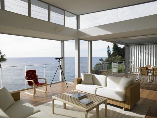 Bright, open-plan coastal living space with floor-to-ceiling glazing overlooking the ocean. The room features a white upholstered sofa and armchair, woven rattan-style furniture, a light timber coffee table, timber decking, and a red lounge chair. A dining area sits on the covered outdoor deck to the right, separated by vertical louvres. High ceilings, clerestory windows, and expansive glazing create an airy indoor-outdoor connection.
