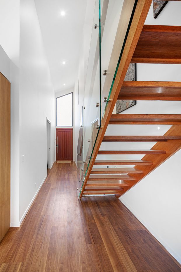 A bright, narrow residential hallway beneath an open-riser timber staircase. The staircase has warm dark-wood treads and structural members, clear glass balustrade panels with metal fixings, and white walls and ceiling with recessed downlights. A timber floor continues through the hall toward a red timber entry door, with a simple contemporary architectural composition.