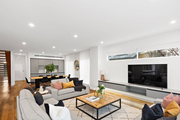Bright contemporary open-plan living and dining area with white walls and recessed ceiling lights. The room features a large sectional sofa arrangement, black-and-white accent cushions, a timber coffee table with black metal legs, a dining table with black chairs, timber flooring, a built-in grey media wall, and extensive horizontal glazing. A hallway and timber louvred door are visible in the background.