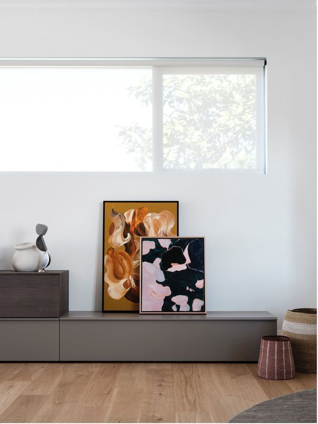 Bright minimalist interior featuring a low, dark grey built-in media console beneath a large translucent roller blind. Abstract artwork leans against the wall alongside a ceramic vase and sculptural ornament, with a woven basket and upholstered stool at right. The room has pale timber flooring, white walls, and soft natural light.