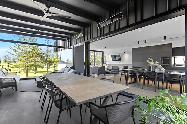 A bright, open-plan contemporary interior combining a dining area, living area and kitchen. A large timber dining table with black-framed chairs occupies the foreground, while the rear features a streamlined dark kitchen with island seating, extensive glazing, exposed dark structural framing and a covered outdoor connection. The space opens onto a landscaped garden and receives abundant natural light.
