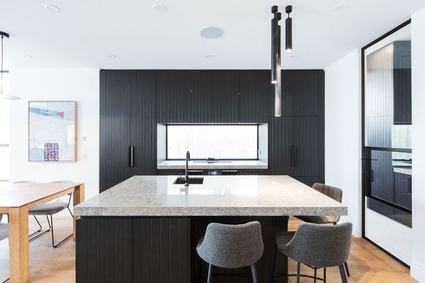 Bright contemporary kitchen with a large light stone island and integrated sink, dark vertically grooved cabinetry, pendant lights, timber flooring, and seating for four. A dining table and artwork are visible to the left, with additional dark joinery and glazing at right.