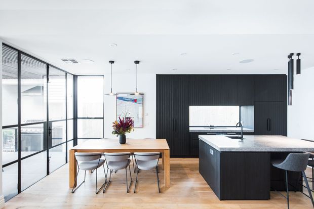 Bright open-plan kitchen and dining area with a light timber dining table and chairs, a large dark vertically grooved kitchen island, black-framed glazed doors and windows, and a matching dark feature wall. Pendant lights hang over the dining table, with generous natural light and pale timber flooring creating a clean contemporary interior.