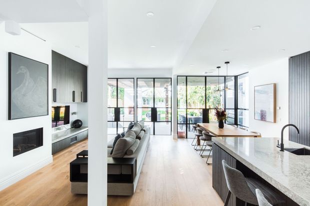 Bright open-plan contemporary interior combining a living room, kitchen and dining area. The space features white walls and ceilings, light timber flooring, dark vertical-panel cabinetry, a pale stone kitchen island, extensive black-framed glazing, minimalist furnishings, and a linear fireplace with large artwork.