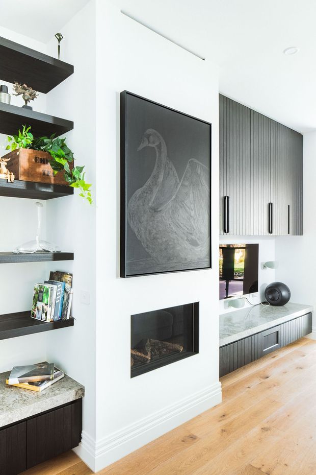 Bright contemporary living room media wall with a large framed artwork, built-in linear fireplace, television, and dark vertically fluted cabinetry. Open dark timber shelving with books, plants, and decorative objects sits to the left, while pale timber flooring and light stone benchtops add warmth and contrast.