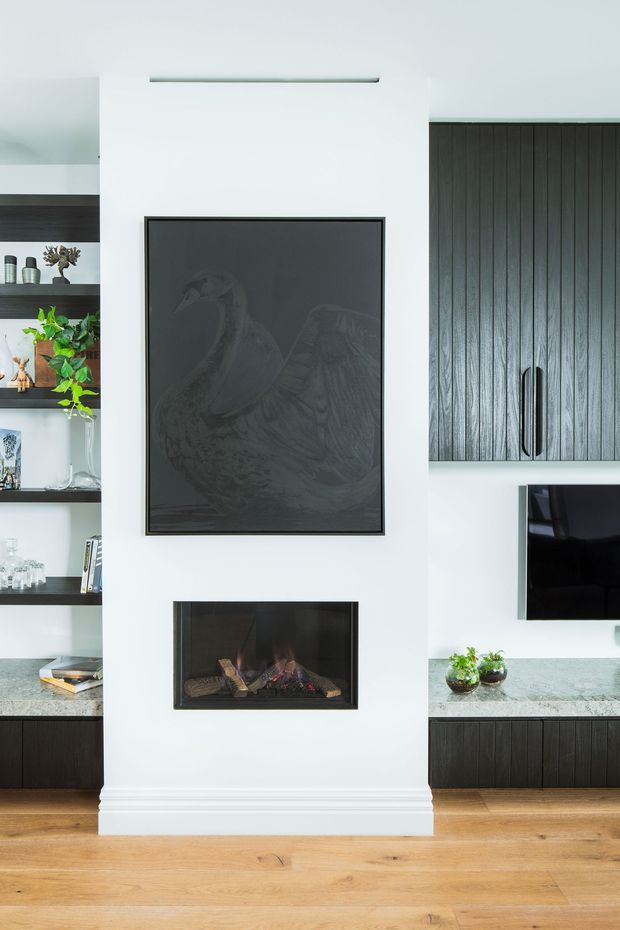 A refined living room media wall with a central white chimney-breast-style feature wall, inset gas fireplace, and dark framed artwork. Built-in shelving and dark vertical-panel cabinetry flank the wall, with a television and pale stone benchtop on the right. Warm timber flooring contrasts with the monochrome joinery and white walls.
