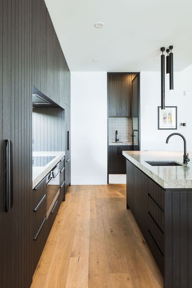 A contemporary galley-style kitchen with dark vertically fluted cabinetry on both sides, a large central island with a pale stone benchtop and integrated sink, warm timber flooring, white walls and ceiling, and black pendant lighting. A compact tiled utility or pantry area is visible through the rear opening.