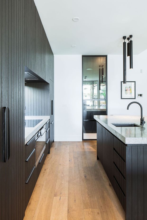 A refined contemporary kitchen viewed down a narrow galley-like passage, with dark vertically fluted cabinetry on both sides, integrated appliances, a large central island with a pale stone benchtop, undermount sink and black gooseneck tap. Warm timber flooring contrasts with white walls and ceiling, while black pendant lights and framed artwork add graphic accents. A glazed internal doorway at the rear reflects additional cabinetry and lighting.