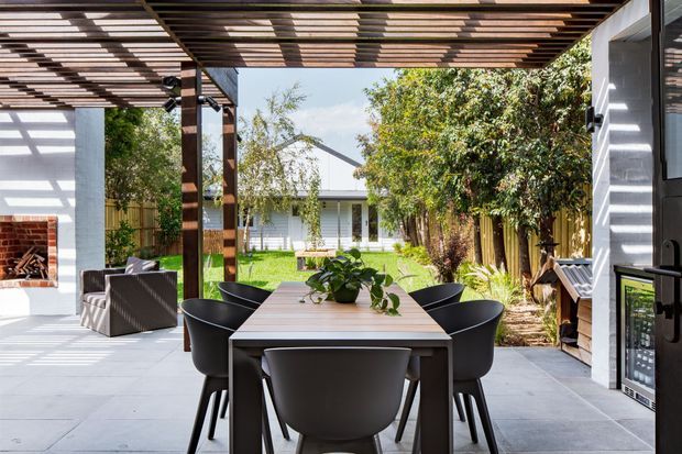 Bright outdoor dining and entertaining area beneath a timber slatted pergola. A long dining table with black chairs sits on a large grey-paved terrace, with an outdoor lounge area, built-in barbecue, and drinks fridge visible around the edges. The space opens onto a landscaped garden and a low white building beyond.