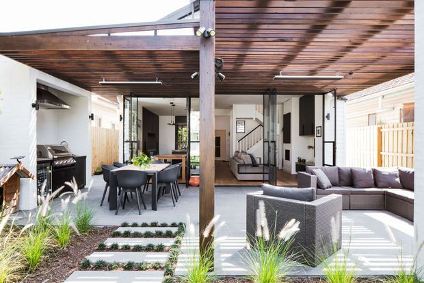 Bright contemporary outdoor entertaining area beneath a timber pergola, with a dining setting, built-in barbecue, outdoor lounge seating, paved surfaces, and landscaped garden beds. The space connects directly to the open-plan interior through large glazed doors.