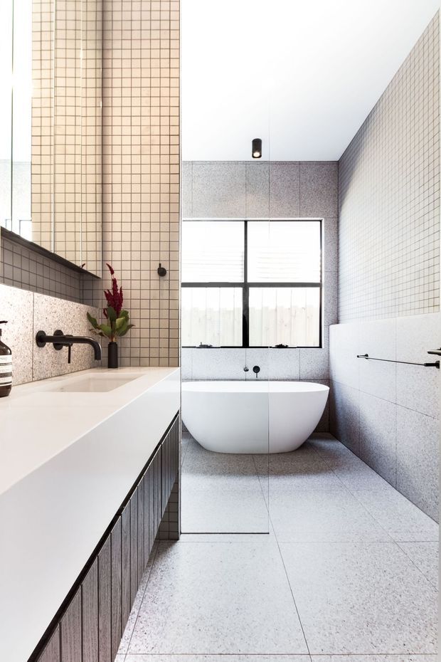 Bright contemporary bathroom featuring a freestanding white bathtub beneath a large window, full-height small-format square wall tiles, pale speckled stone or terrazzo-style floor and wall surfaces, and a long white vanity with dark ribbed cabinetry and matte black fixtures. A slim pendant light and restrained detailing create a clean, refined atmosphere.