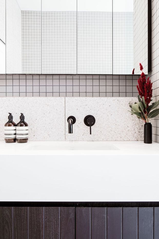 A close-up view of a contemporary bathroom vanity featuring a large white rectangular basin, matte black wall-mounted tapware, pale terrazzo-like wall panels, and small square grid tiles in white and charcoal grey. A frameless mirrored cabinet spans the upper wall, with dark soap dispensers and a black vase holding dried red foliage on the counter. The palette is monochrome with deep charcoal accents and a clean, refined finish.