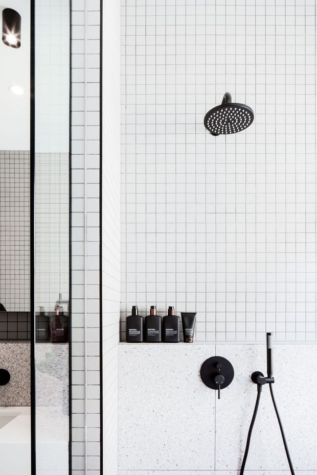 Close-up of a contemporary bathroom shower with white square wall tiles, dark grout lines, matte black shower fittings and controls, and a pale speckled stone or terrazzo wall panel. A frameless glass shower screen reflects dark bathroom joinery and toiletries at the left.