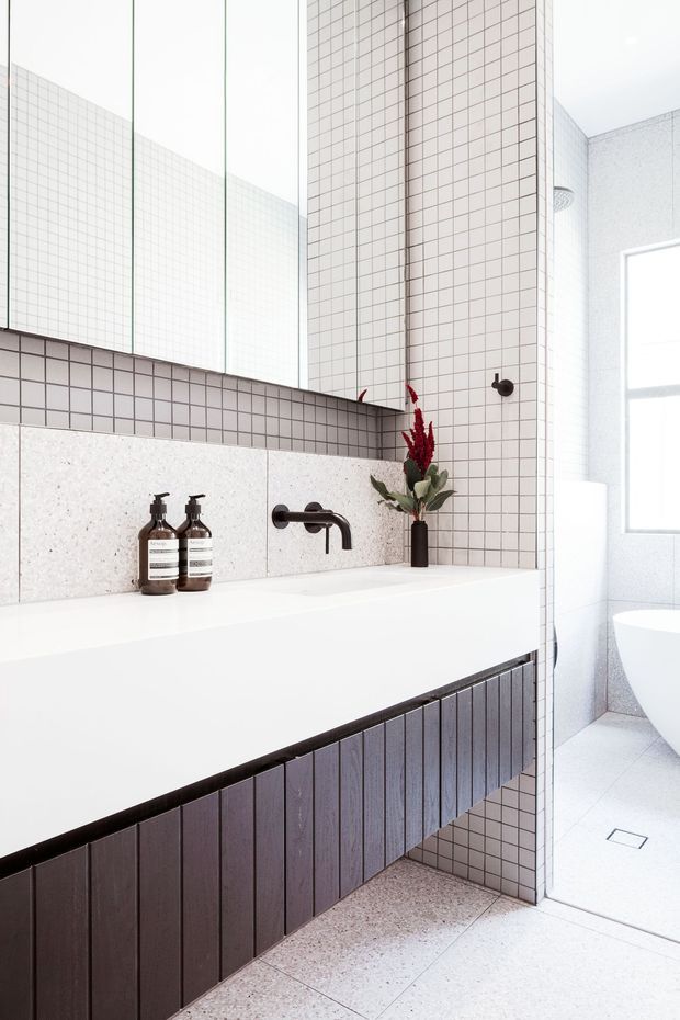 Bright contemporary bathroom with a long white floating vanity, dark vertically slatted timber cabinetry, pale square wall tiles, terrazzo-like wall and floor surfaces, and matte black wall-mounted tapware. A large mirror spans the vanity, while a shower and freestanding bathtub are visible through the opening at right. Minimal styling includes dark soap dispensers and a small vase with red foliage.