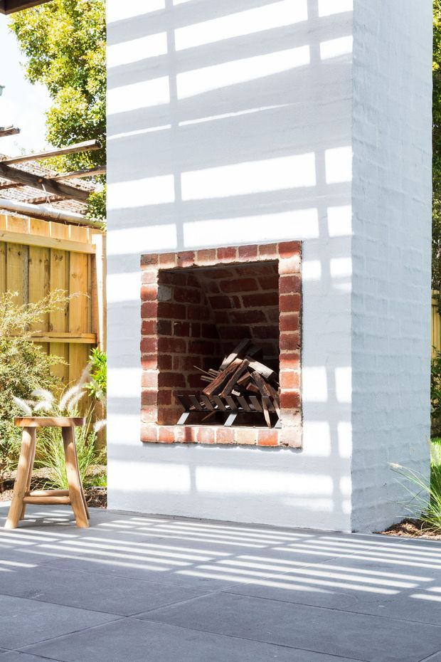 Bright outdoor patio area featuring a white-painted brick fireplace with a red brick firebox and stacked firewood. A timber privacy fence, garden planting, timber stool, and strong linear shadows from overhead screening or pergola structure surround the fireplace.