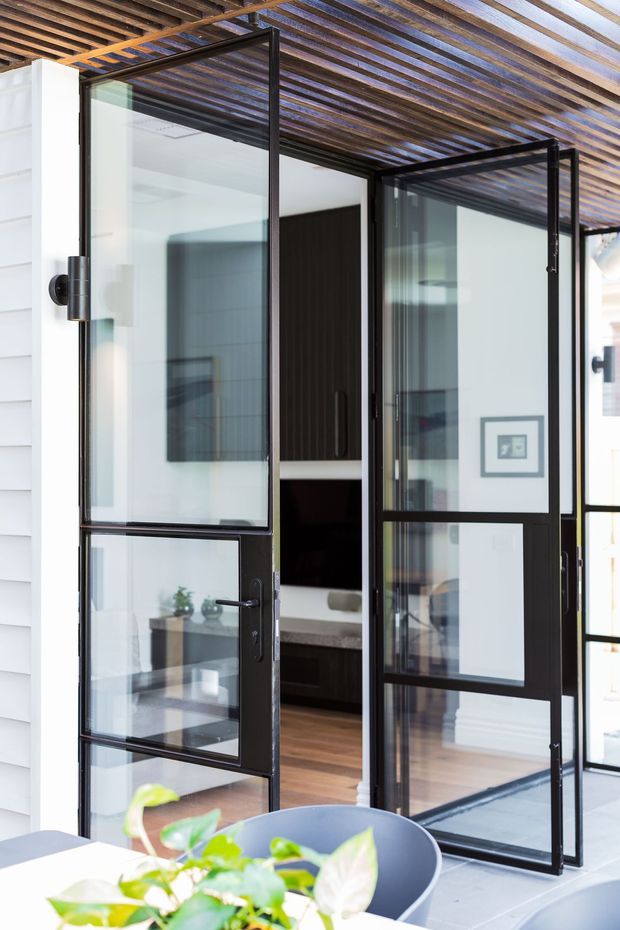 A bright contemporary indoor-outdoor threshold with black-framed glazed pivot or hinged doors opening onto a covered terrace. The doors feature multiple glass panels and slim dark metal frames, with a timber slatted pergola overhead. White horizontal cladding is visible at the exterior, while a modern interior with dark cabinetry, timber flooring, and framed artwork can be seen beyond the doorway. A dining chair and leafy plant appear in the foreground.