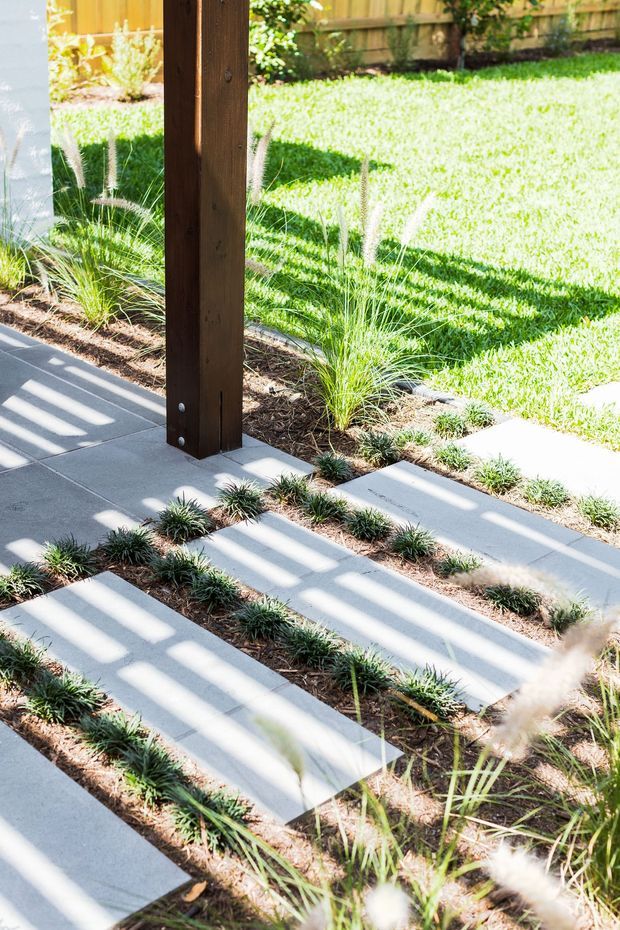 A bright landscaped outdoor walkway or patio area with large pale concrete stepping slabs arranged in parallel rows through planted garden beds. Ornamental grasses and low groundcover surround the path, while a dark timber structural post stands prominently near the edge. Strong sunlight creates linear shadows across the paving and lawn, with a timber boundary fence visible in the background.