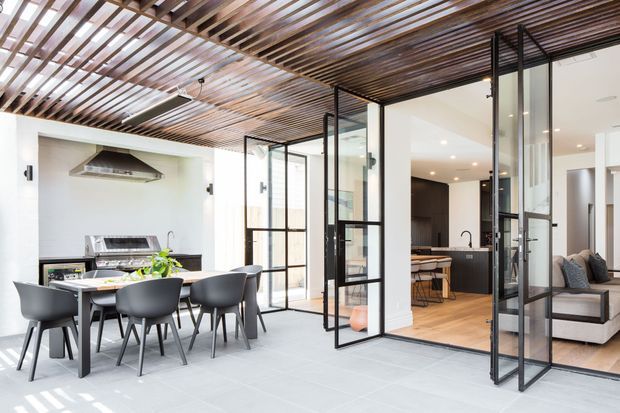 Bright contemporary open-plan indoor-outdoor living and dining area with a large dining table, modern chairs, an outdoor-style kitchen/barbecue zone, and black-framed glazed folding doors opening into a warm timber-floored kitchen and living room. A slatted timber ceiling creates a strong architectural feature across the space.