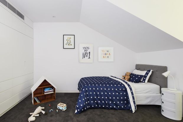 A bright, compact children's bedroom with a single bed, navy star-patterned bedding, a grey upholstered headboard, white built-in wardrobes, dark carpet, a small white bedside table, and playful toys and artwork. The sloped ceiling creates a cosy attic-like feel.