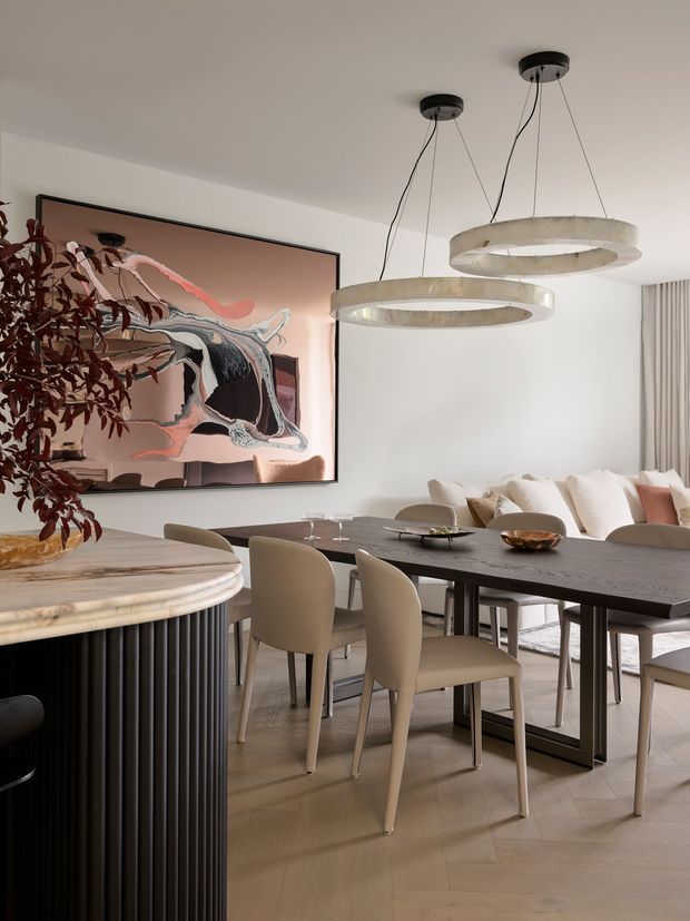 Refined contemporary dining and living area with a dark rectangular dining table, upholstered cream dining chairs, sculptural circular pendant lights, and a pale upholstered sofa. A large abstract artwork provides a muted blush, black, and cream focal point, while a curved stone-topped island or console with dark fluted joinery occupies the foreground. The room has light timber flooring, white walls, and soft neutral finishes.