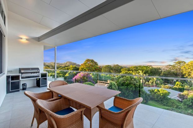 Covered outdoor balcony or terrace with a dining table and woven wicker chairs, built-in barbecue cabinetry along the wall, tiled flooring, glass balustrade, and a broad leafy suburban outlook. The space is open-sided and designed for outdoor dining and entertaining.