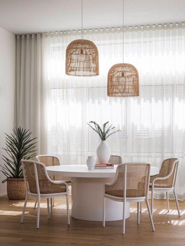 Bright, airy dining room with a round white pedestal dining table surrounded by woven cane dining chairs. Floor-to-ceiling sheer curtains filter abundant daylight, while two suspended rattan pendant lights and a tall potted plant add natural texture. The room has warm timber flooring and a soft neutral palette.